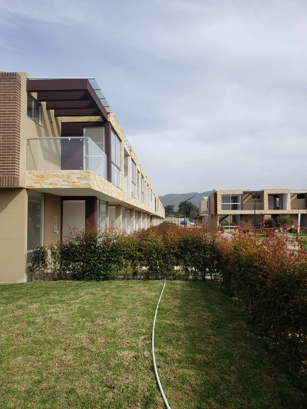 Foto real de jardines laterales de Bosco Reservado con setos rojos y verdes y casas en construcción al fondo